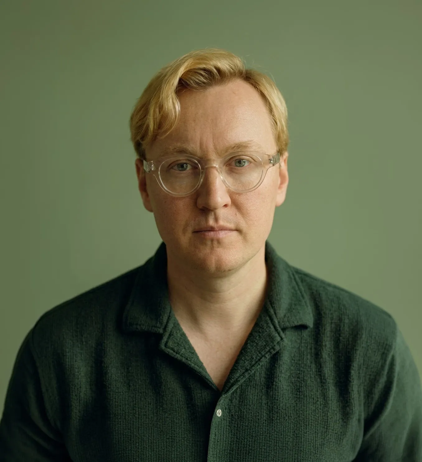 Green-toned editorial studio portrait of a man wearing clear round glasses, featured on the WeGrow site.